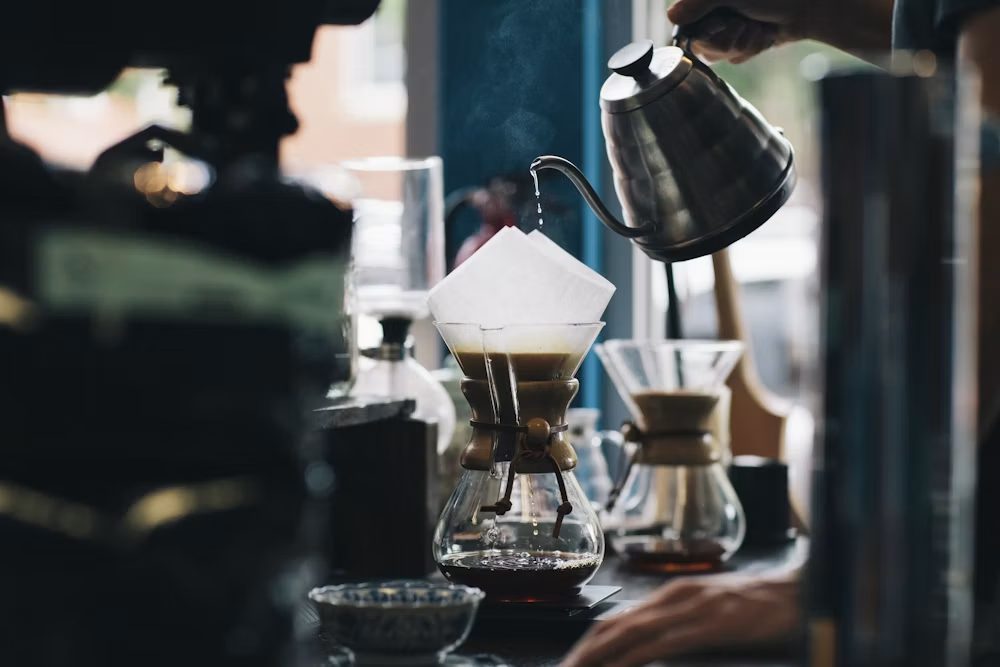 A barista pouring from a gooseneck kettle into a Chemex pour-over by a window